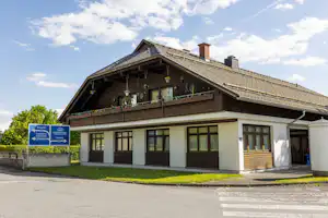 Campinghaus mit Holzveranda und Balkon, Fensterfront und Schornstein, davor asphaltierter Weg und Beschilderung am Eingang.