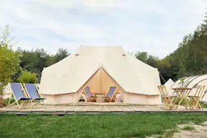 Großes Zelt auf einer Campingwiese mit Sonnenliegen und Holzstühlen, im Hintergrund Bäume und ein Weg.
