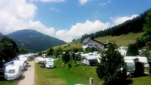 Campingplatz mit Wohnwagen und Wohnmobilen auf grüner Wiese, im Hintergrund Berge und ein Gebäude am Hang.