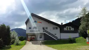 Campinggebäude mit „alpenwelt“-Schriftzug, Treppe und Balkon, davor gepflasterter Weg, Rasen und Bäume vor bewölktem Himmel.