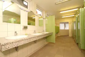 Gemeinschaftswaschraum mit zwei Waschbecken, Fliesenboden, Spiegeln und grünen Spinden sowie Handtuchhaltern an der Wand.