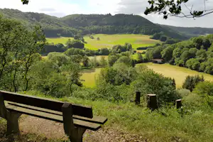 Blick von einer Bank auf grüne Wiesen, bewaldete Hügel und ein kleines Holzhaus im Tal.