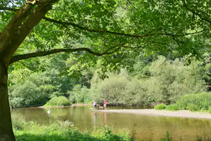 See am Campingplatz unter großen Bäumen, im Hintergrund zwei Personen mit Hunden am Ufer.