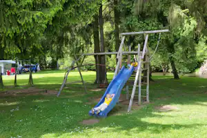 Blauer Spielplatz-Rutschenturm mit zwei Kindern auf dem Campingplatz im Grünen, neben Bäumen und einem kleinen See.