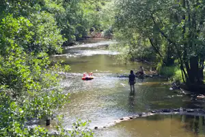 Flussufer im Grünen mit Raftboot im Wasser, Personen am Ufer und Bäume im Hintergrund.