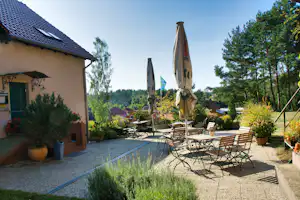 Terrasse am Campingplatz mit Tisch und Stühlen, zwei Sonnenschirmen, Pflanzenkübeln und Blick in den Garten mit Bäumen.