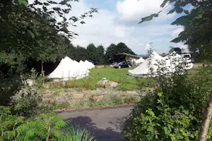Zeltplatz im Grünen: zwei weiße Zelte auf einer Wiese, dazwischen Steine, Wege und Büsche im Vordergrund.