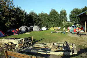 Campingwiese mit mehreren Zelten, Sitzgruppe und Holztisch unter Bäumen, im Vordergrund eine Feuerstelle.