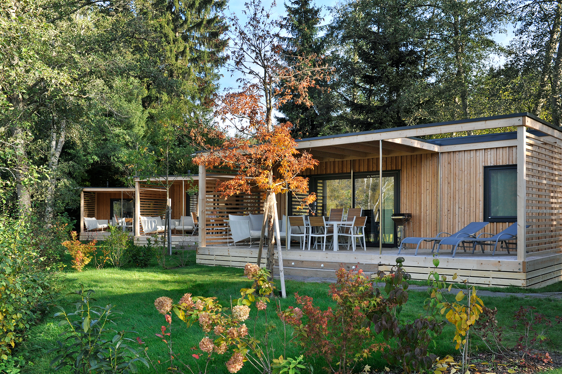 Holzunterkunft mit Terrasse, Liegestühlen und Glasfront im Garten, umgeben von Bäumen und Blumenbeeten.