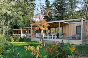 Holzunterkunft mit Terrasse, Liegestühlen und Glasfront im Garten, umgeben von Bäumen und Blumenbeeten.