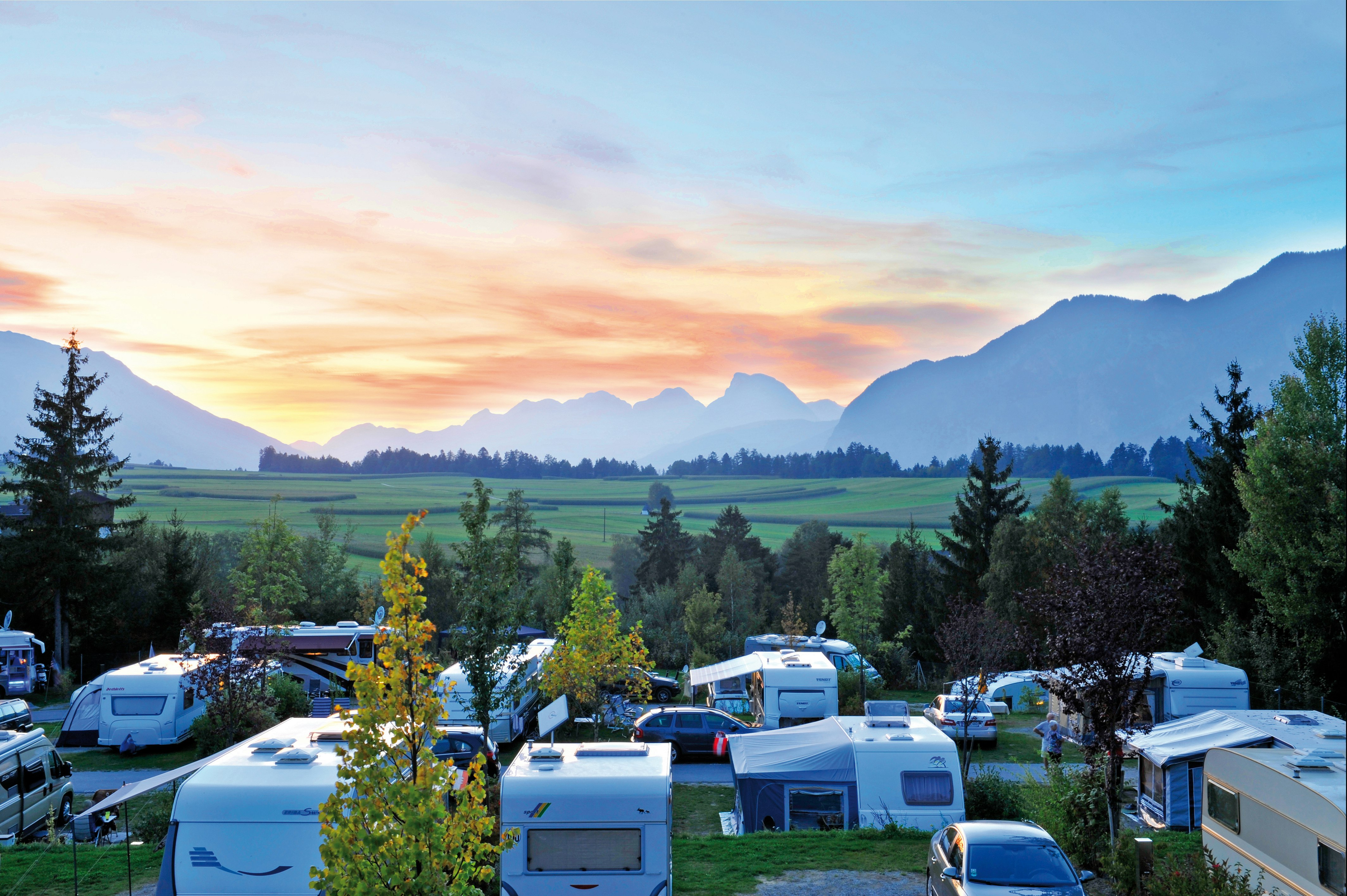 Campingplatz mit Wohnwagen und Stellplätzen, Blick ins Tal mit Wiesen, Bäumen und Bergen in der Ferne.