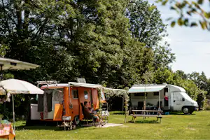 Wohnwagen und Zelt auf einem grünen Campingplatz, mit Sonnenschirm, Stühlen und Tisch vor dem Wohnwagen.