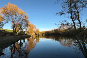Ruhiger Campingsee mit Spiegelung von Herbstbäumen und Uferbäumen unter klarem Himmel.