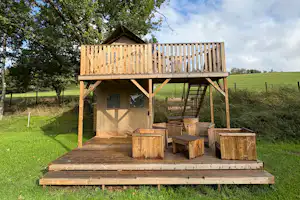 Holzspielhaus auf einer Wiese mit Treppe, Holzboxen und Veranda unter hohen Bäumen.