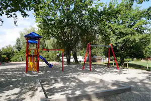 Buntes Spielgerät mit Rutsche und Schaukeln auf Kiesfläche, umgeben von hohen Bäumen und Wohnwagen im Hintergrund.