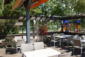 Terrasse mit Tisch und Stühlen, Blick auf den Teich, Blumenbeet und Spielbereich mit bunten Zelten im Grünen.