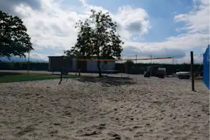 Sandiger Beach-Volleyballplatz mit Netz und Zaun, im Hintergrund Bäume und ein Gebäude am Campinggelände.