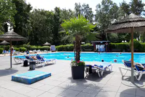 Camping-Pool mit blauen Liegen, Palmen und Sonnenschirmen, im Hintergrund ein Gebäude und Hecken im Garten.