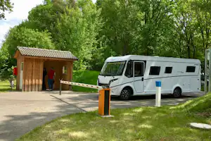 Wohnmobil auf dem Stellplatz neben einem kleinen Holzhaus, mit Schranke und Bäumen im Hintergrund.