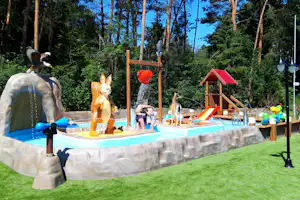 Wasserspielplatz mit Tierfiguren im Pool, Rutsche und Spritzwasser, umgeben von Bäumen und Rasenfläche.