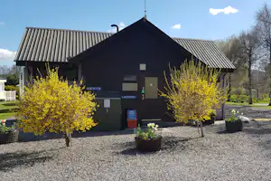 Campinggebäude mit Eingangstür, Schildern und Treppe, davor Kiesweg und zwei gelbe Bäume in Kübeln.
