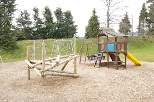 Spielplatz am Campingplatz mit Klettergerüst, Rutsche und Schaukeln auf einem Kiesplatz, umgeben von hohen Bäumen.