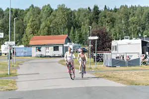 Zwei Radfahrer auf dem Weg am Campingplatz, Wohnwagen und kleine Gebäude im Hintergrund, Bäume und Laternen entlang der Straße.