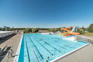 Großes Camping-Schwimmbecken mit blauen Bahnen und orangefarbener Rutsche, daneben Wege und Liegeflächen am Beckenrand.