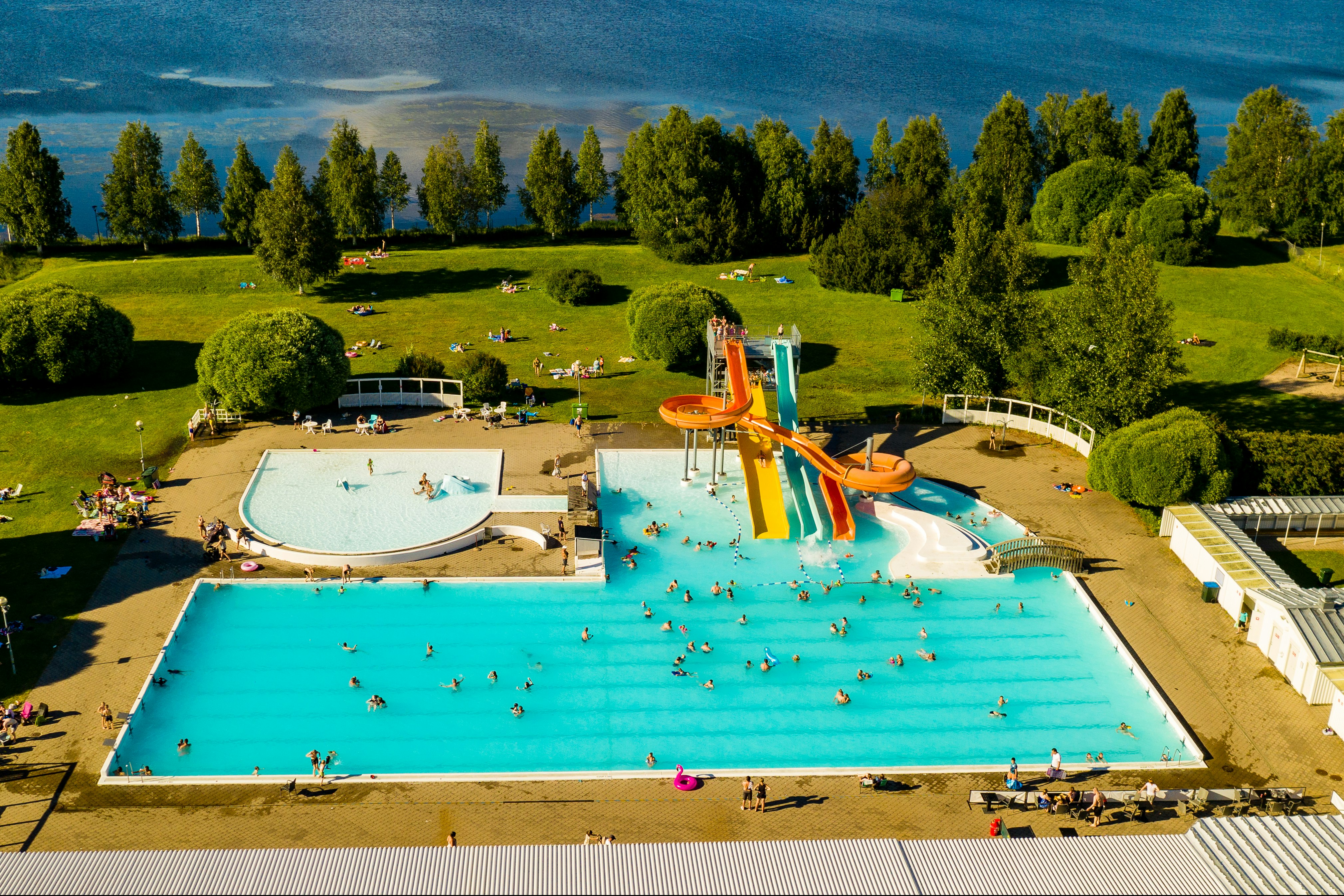 Großes Schwimmbecken mit mehreren Bahnen und Rutschen, umgeben von Liegen, Rasenflächen und Bäumen auf einem Campingplatz.