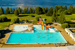 Großes Schwimmbecken mit mehreren Bahnen und Rutschen, umgeben von Liegen, Rasenflächen und Bäumen auf einem Campingplatz.