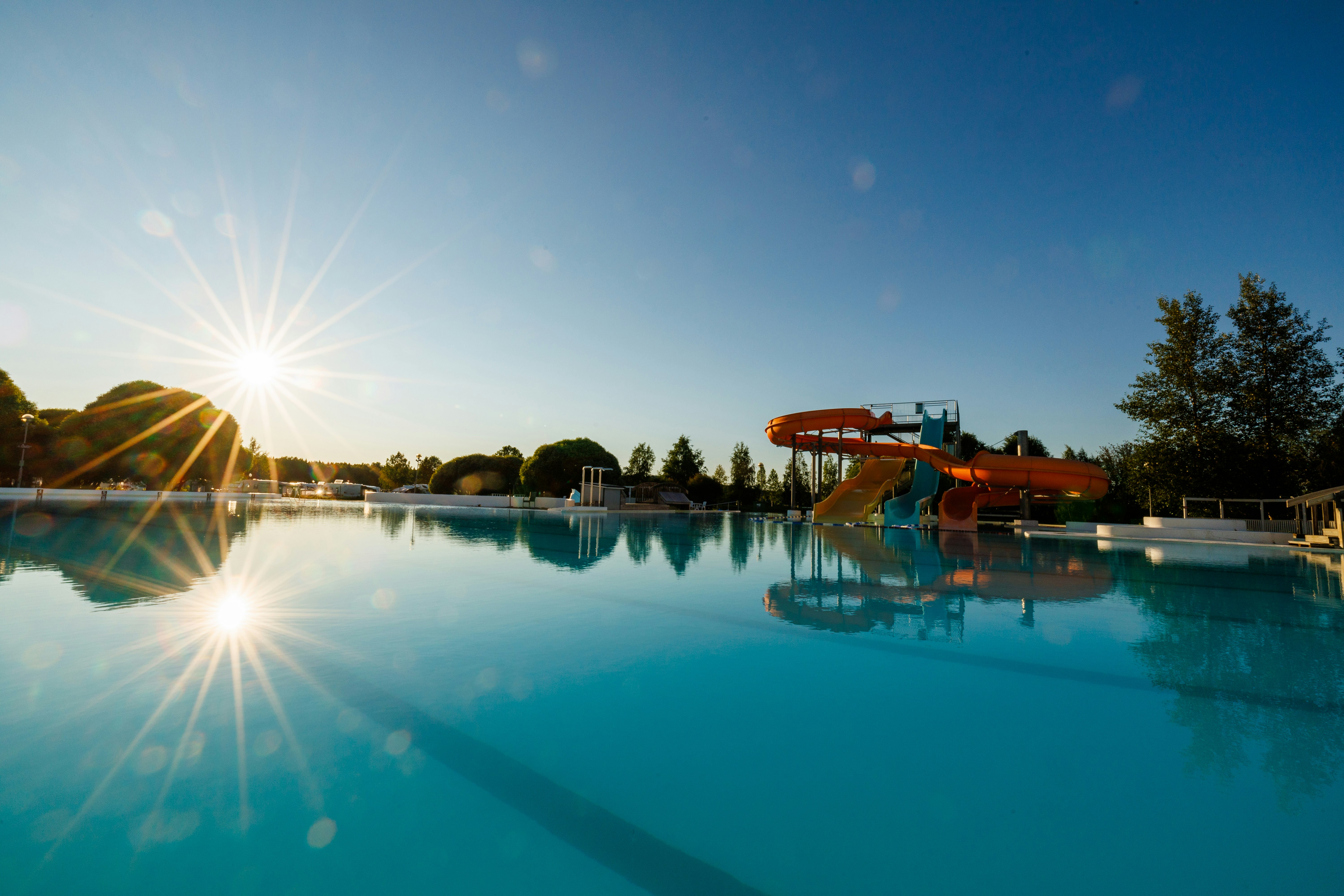 Schwimmbecken im Campingbereich mit Rutsche, ruhiges Wasser und Sonnenreflexen, im Hintergrund Bäume und Gebäude.