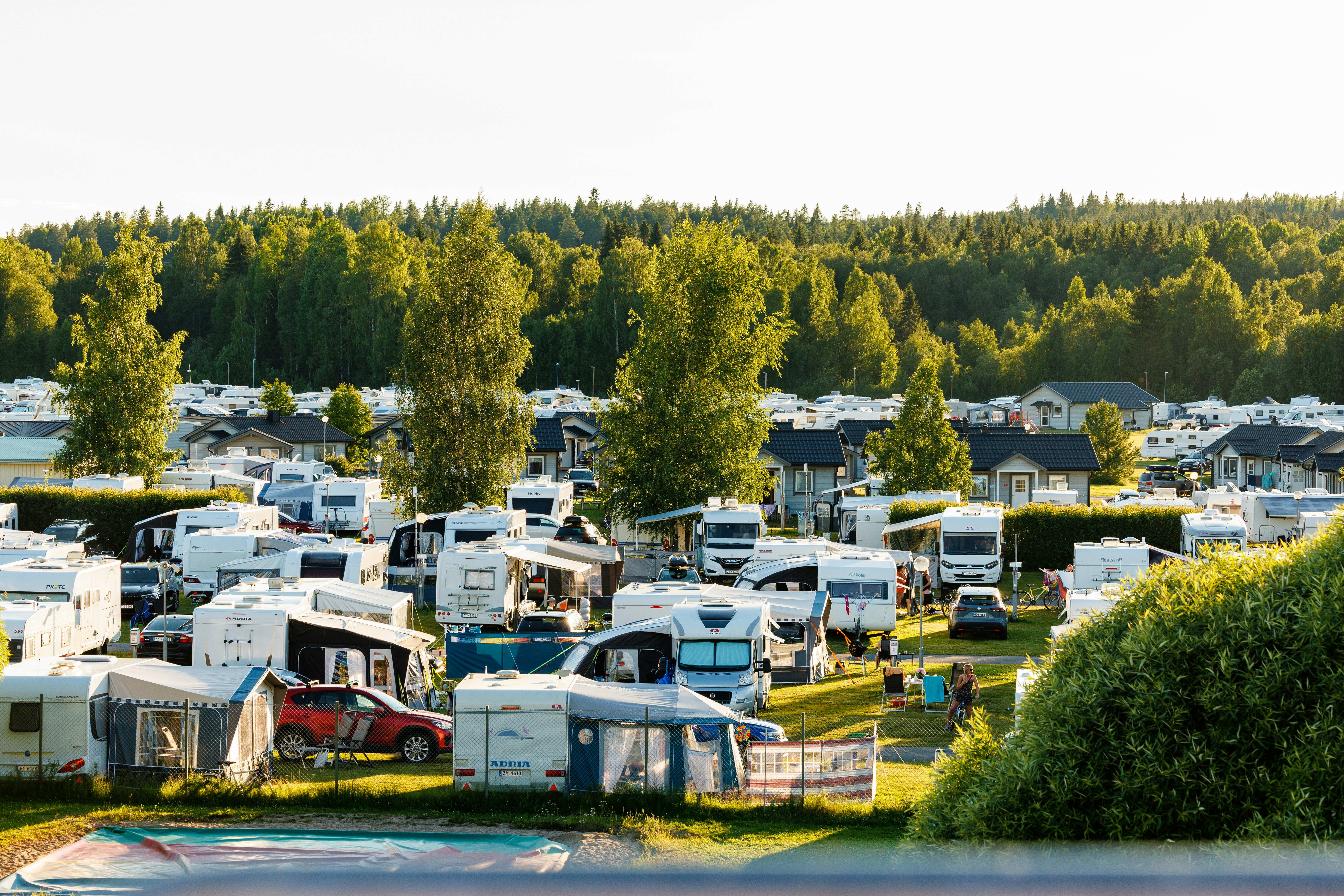Campingplatz mit Wohnmobilen und Zelten, Stellplätzen im Grünen, Bäumen und Häusern am Seeufer im Hintergrund.