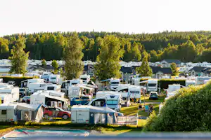 Campingplatz mit Wohnmobilen und Zelten, Stellplätzen im Grünen, Bäumen und Häusern am Seeufer im Hintergrund.
