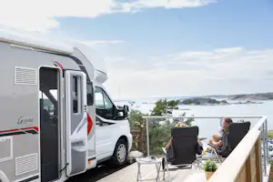 Wohnmobil mit geöffneter Tür und Terrasse mit Stühlen am See, im Hintergrund Wasser und Uferlandschaft.