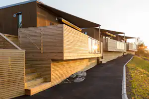 Holzterrasse an einer Campingunterkunft mit Treppe, Geländer und beleuchtetem Eingang, daneben ein gepflasterter Weg.