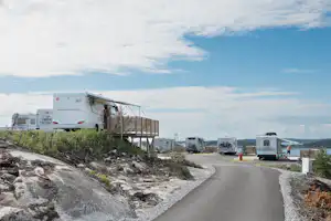 Wohnmobile auf einem Campingplatz, Holzplattformen und Schotterwege, im Hintergrund weitere Fahrzeuge und ein Seeufer.