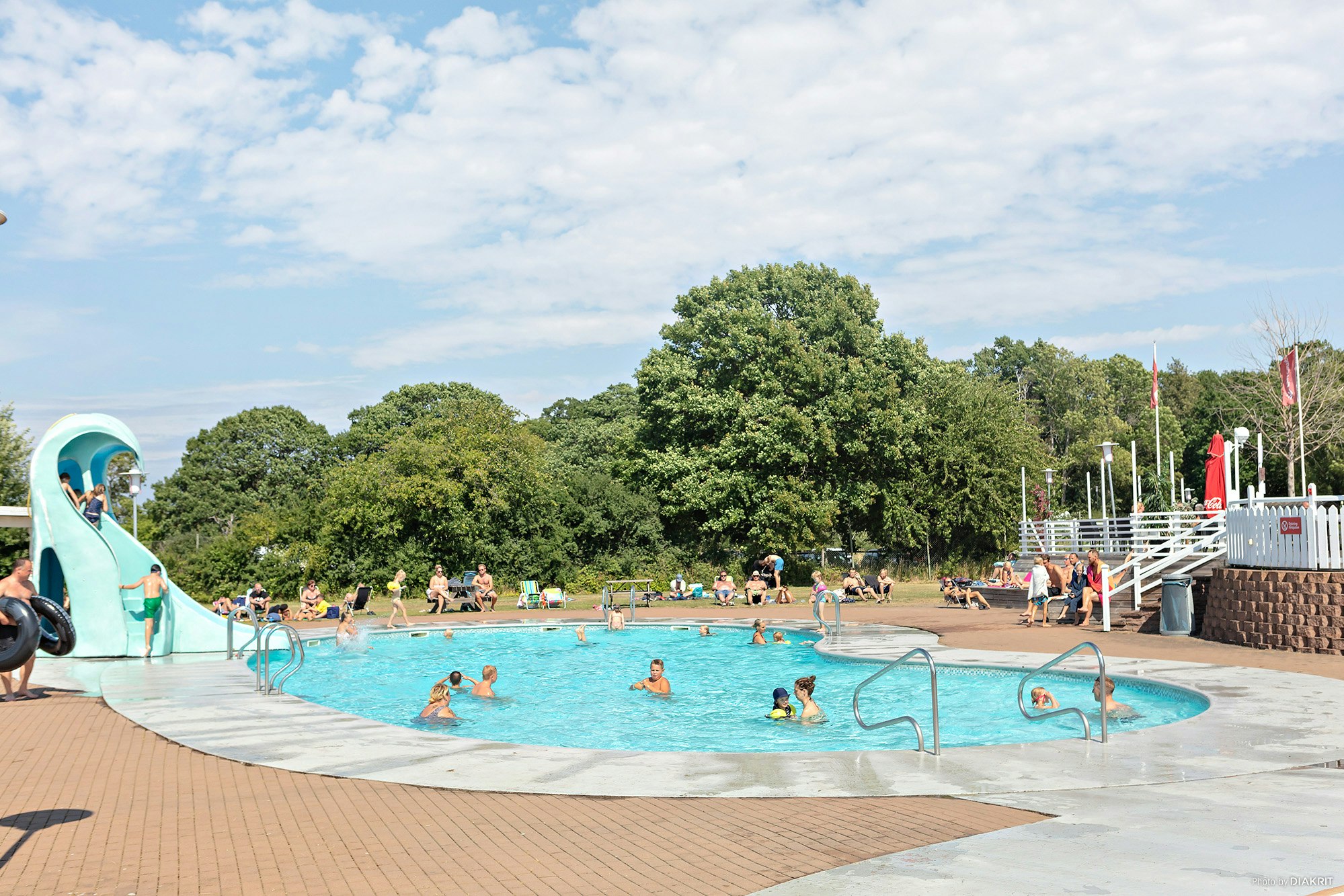 Kinderbereich am Campingplatz mit Wasserrutsche, Schwimmbecken und Liegen am Rand, im Hintergrund Bäume und weitere Gäste.