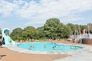 Kinderbereich am Campingplatz mit Wasserrutsche, Schwimmbecken und Liegen am Rand, im Hintergrund Bäume und weitere Gäste.