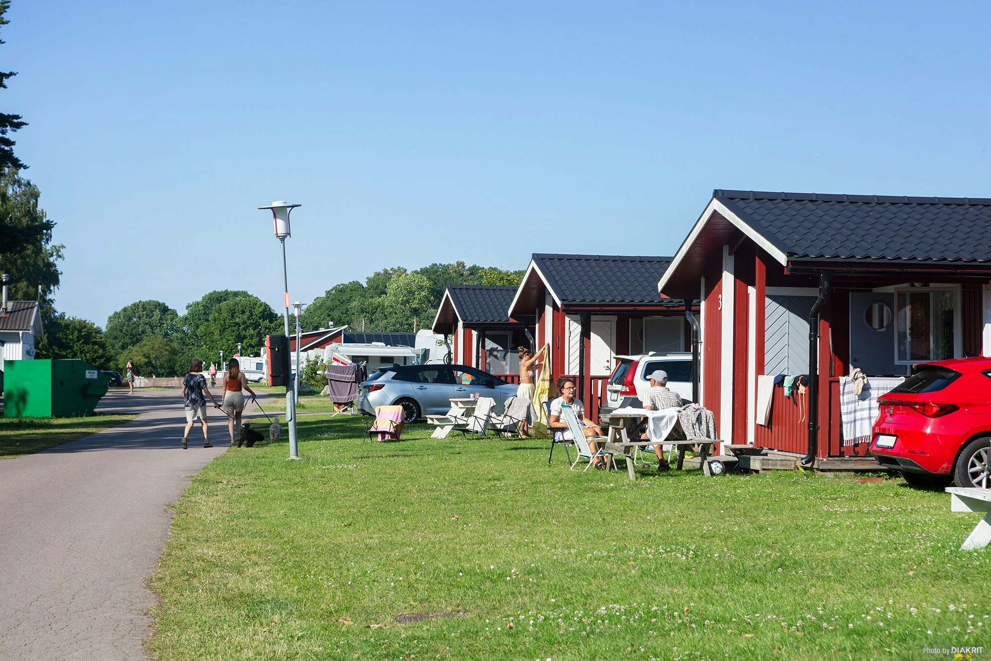 Reihen von Campinghäusern mit roten Fassaden, Rasenfläche und Sitzgruppen; im Vordergrund ein roter Pkw und Wohnwagenanhänger.