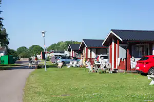 Reihen von Campinghäusern mit roten Fassaden, Rasenfläche und Sitzgruppen; im Vordergrund ein roter Pkw und Wohnwagenanhänger.