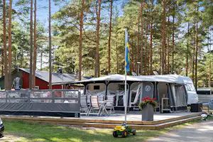 Wohnwagen mit Vorzelt und Terrasse auf einem Stellplatz im Wald, daneben Gartenmöbel und ein kleiner Spielzeugtraktor.