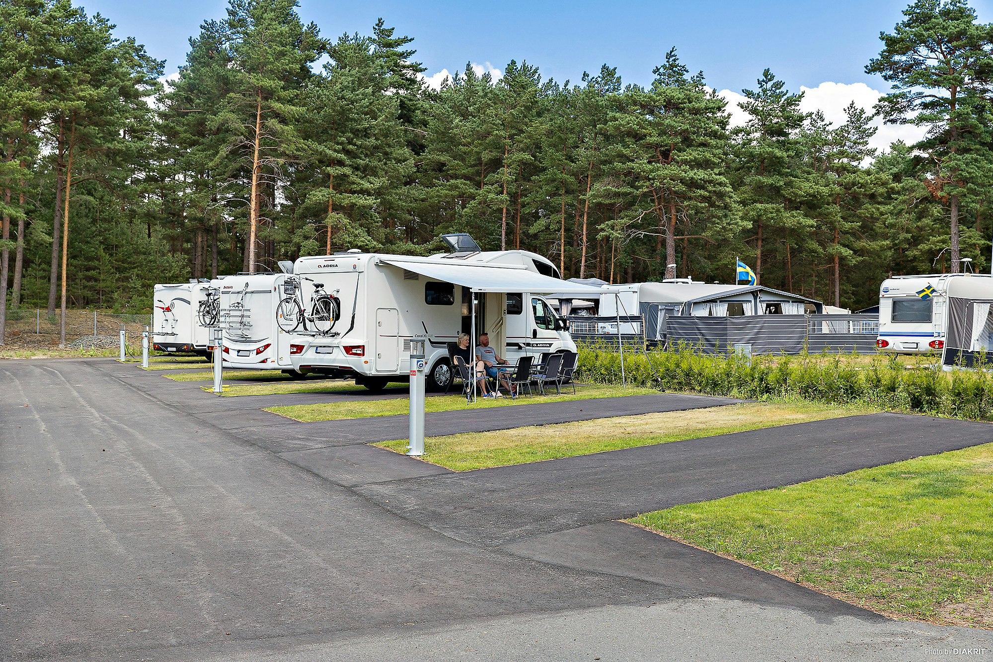 Wohnmobilstellplätze im Wald mit mehreren Caravans und einem weißen Wohnmobil, daneben Tisch und Stühle auf dem Betonplatz.