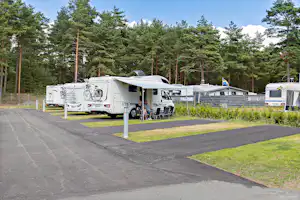 Wohnmobilstellplätze im Wald mit mehreren Caravans und einem weißen Wohnmobil, daneben Tisch und Stühle auf dem Betonplatz.