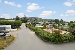 Campingplatz-Gasse mit Wohnwagen und Zelten, Heckenbeete, Spielplatzschaukel und Häusern am Rand, Blick in die Ferne.
