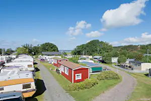 Reihe von Wohnwagen und ein rotes Ferienhaus auf einem Campingplatz, mit Wegen, Hecken und Blick auf Bäume und Wasser.
