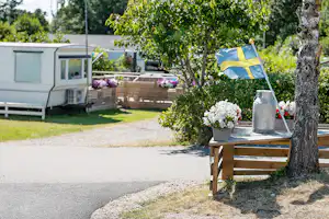 Vor einem Mobilheim stehen eine Holzbank und ein Tisch mit Blumen, daneben eine schwedische Flagge im Garten.