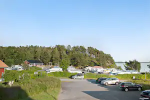 Campingplatz-Parkplatz mit Autos, Hecken und Bäumen, im Hintergrund Gebäude und ein See unter klarem Himmel.