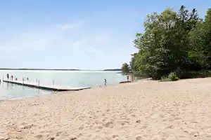 Sandstrand am See mit Steg, Bäumen am Ufer und einigen Personen im Wasser und am Strand.