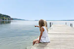 Ein Kind sitzt auf einem Holzsteg am See und schaut ins Wasser, im Hintergrund Ufer und Bojen.