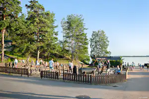 Spielplatz am Campingplatz mit Holzzaun, Rutsche und vielen Kindern am Weg entlang, im Hintergrund Bäume und See.